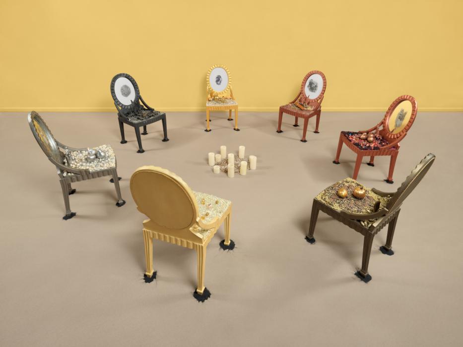 Seven chairs are organized in a circle facing in towards an arrangement of candles. Each is painted a different color. They are butter yellow, metallic grey, black, gold, clay red, and a brown bronze. Each has a rounded back with an image at the center, and beads and other objects rest on their seats. They are on a taupe colored floor and there is a solid butter yellow wall behind them.