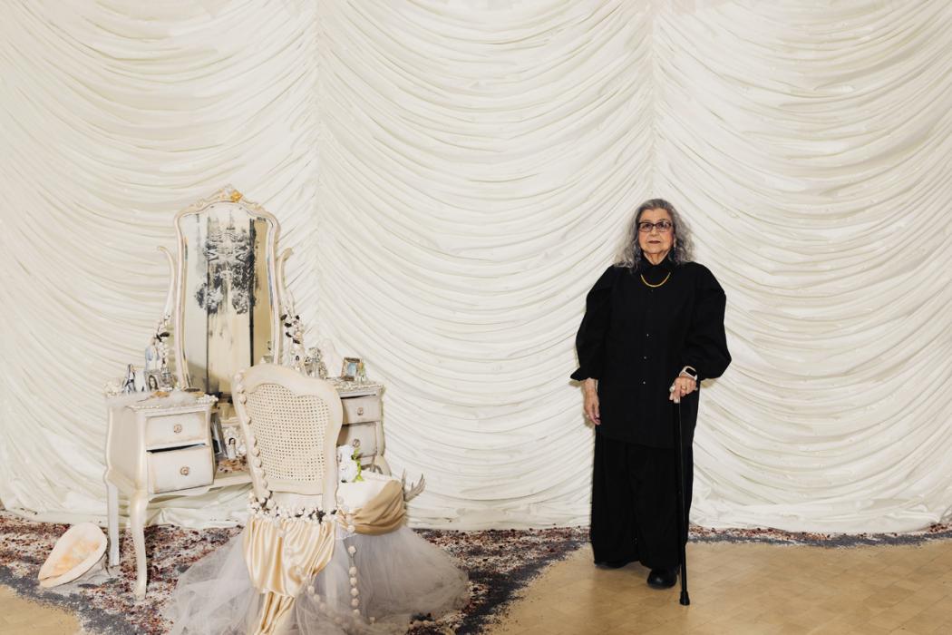 A woman with white grey hair and glasses stands resting one hand on a cane. She wears an all black shirt and skirt. She stands to the right of a work of art, comprising a white dresser and chair, covered in small objects. Behind her is a wall draped with white curtains.
