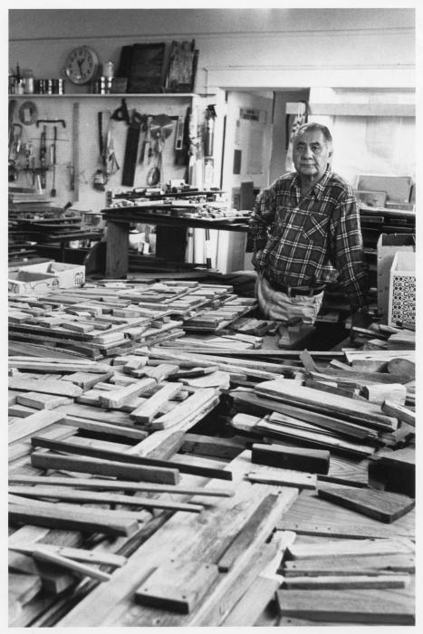 A black and white photograph shows a man with long greying hair standing behind a table filled with pieces of wood of various sizes. He rests his right hand on the table and his left on his hip. He looks directly at the camera with a neutral expression. Behind him is another higher table with pieces of wood. And on the wall behind it are tools for a wood workshop.