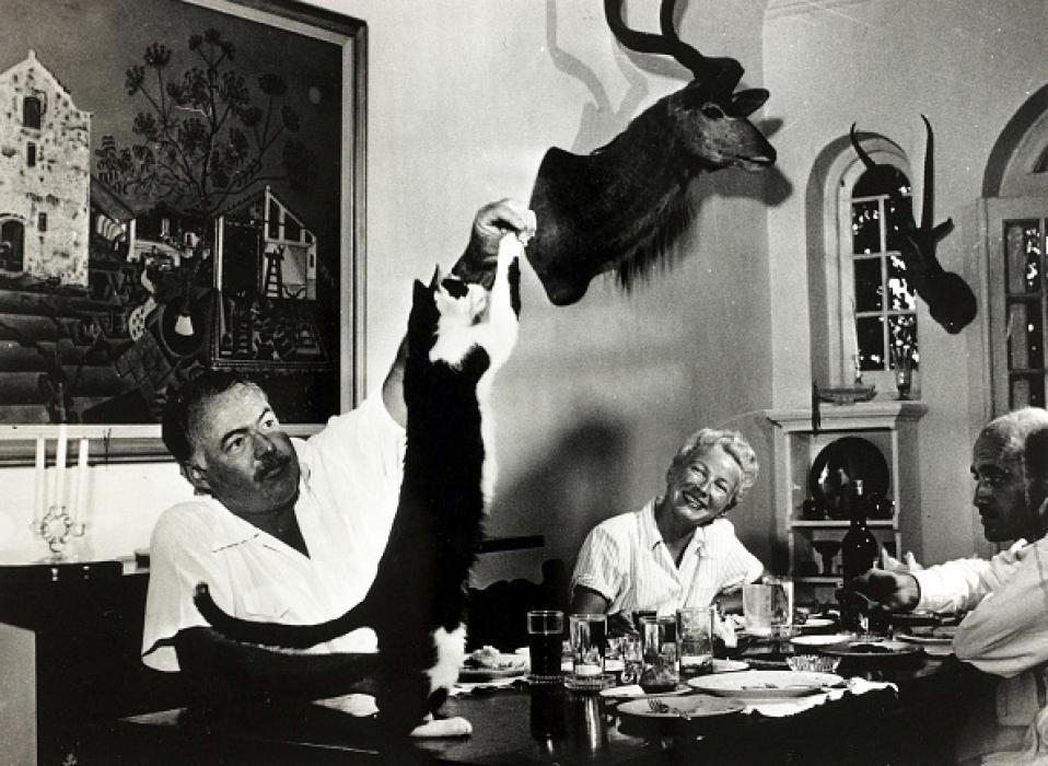 A black and white photo shows a bearded man feeding a cat standing on a table. A woman sits past him at the table. The head of a large antlered animal is on the wall.