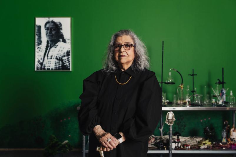 A woman with white grey hair and black glasses stands with her hands clasped at her waist. She is shown from the waist up. Behind her is a bright green wall, with a black and white photograph of a woman to the left. On her right is a metal table of various scientific tools and other small objects.