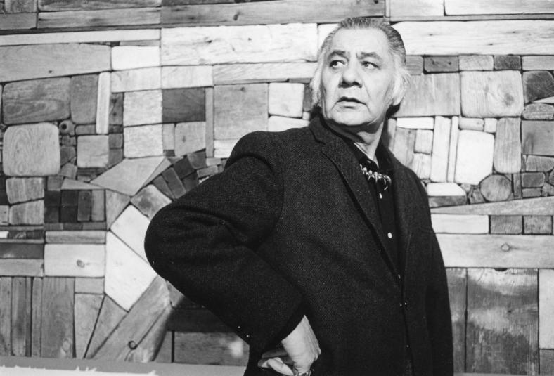 A black and white photograph shows a man with greyish hair standing against a wall of stacked cubes of wood in different shades. He is shown from the waist up. He is turned to the right and looks to the left. He rests his left hand on his waist, with his elbow bent. He has a serious expression.