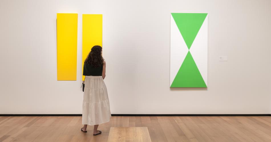 A woman is shown from behind in a white dress with a black sweater hanging over her shoulders. She stands looking towards two abstract paintings hanging on a white wall. A two piece vibrant yellow painting hangs to the left. On the right is a painting with two triangles of kelly green one of which is inverted so they meet in the middle of the canvas. The remaining left and right of the canvas is a solid white.