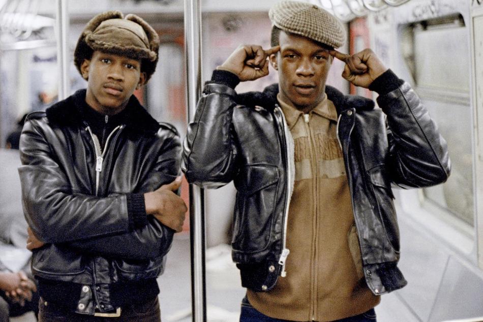 Two Black men stand in a subway car facing the camera. Both are shown from the hips up. The man on the left wears a black leather jacket and brown fur hat, and wraps his arms around the body. He has a neutral expression but direct gaze. The man on the right wears a black leather jacket over a brown sweater. He points a finger on each hand at his head, which is topped by a brown newsboy cap.