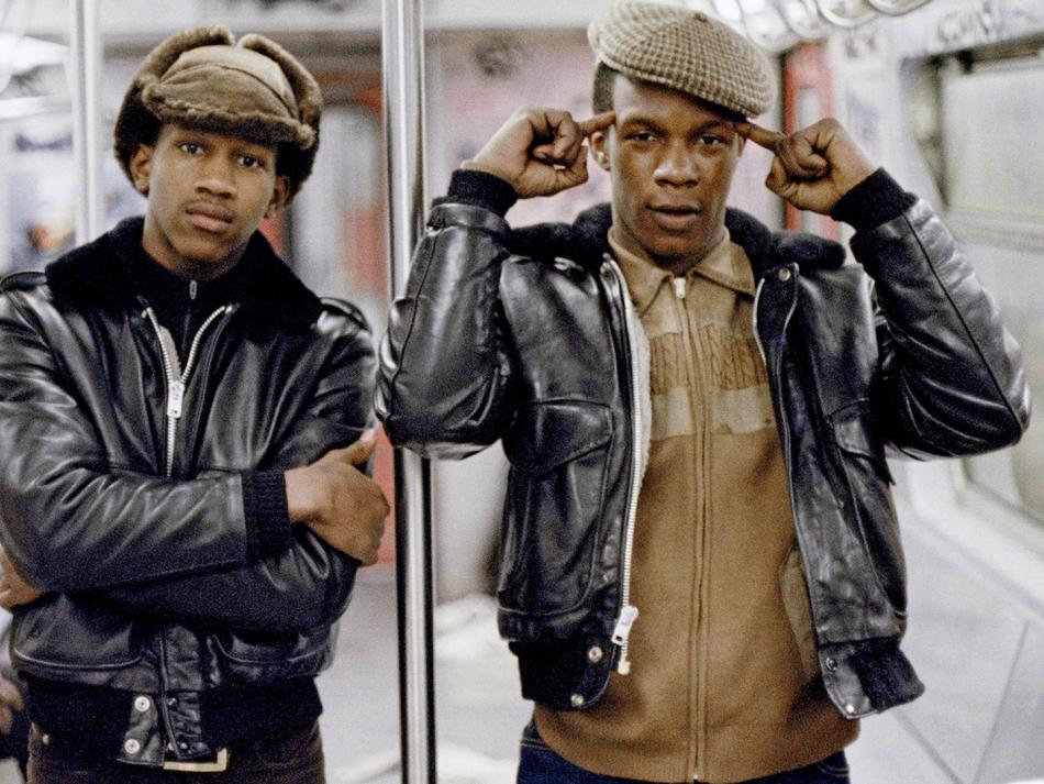 Two Black men stand in a subway car facing the camera. Both are shown from the hips up. The man on the left wears a black leather jacket and brown fur hat, and wraps his arms around the body. He has a neutral expression but direct gaze. The man on the right wears a black leather jacket over a brown sweater. He points a finger on each hand at his head, which is topped by a brown newsboy cap.