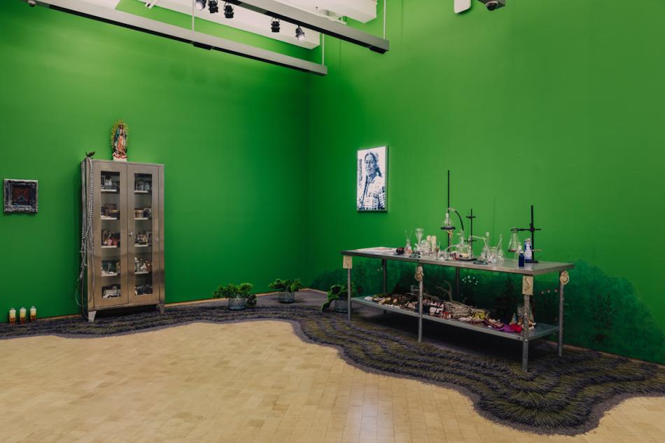 An installation in the corner of a room with bright kelly green walls. On the right is a metal table covered in small objects and scientific tools like test tubes. On the ground in front of it is a curving layer of dried lavender. To the left is a metal cabinet with glass doors. Objects can be seen within it. A sculpture of a woman rests on top of it.