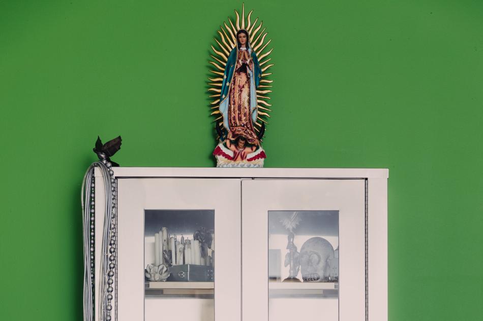 A metal cabinet with glass doors. Objects can be seen in the shelves inside including a skull and books. A sculpture of a woman rests on top of it.
