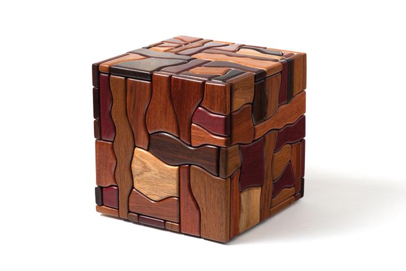 A cube made of pieces of wood in different shapes and stains, from dark brown, to reddish brown, to a light tan color. The cube is set against a white background and shown slightly turned so one corner is facing us. We can see the top and two sides.