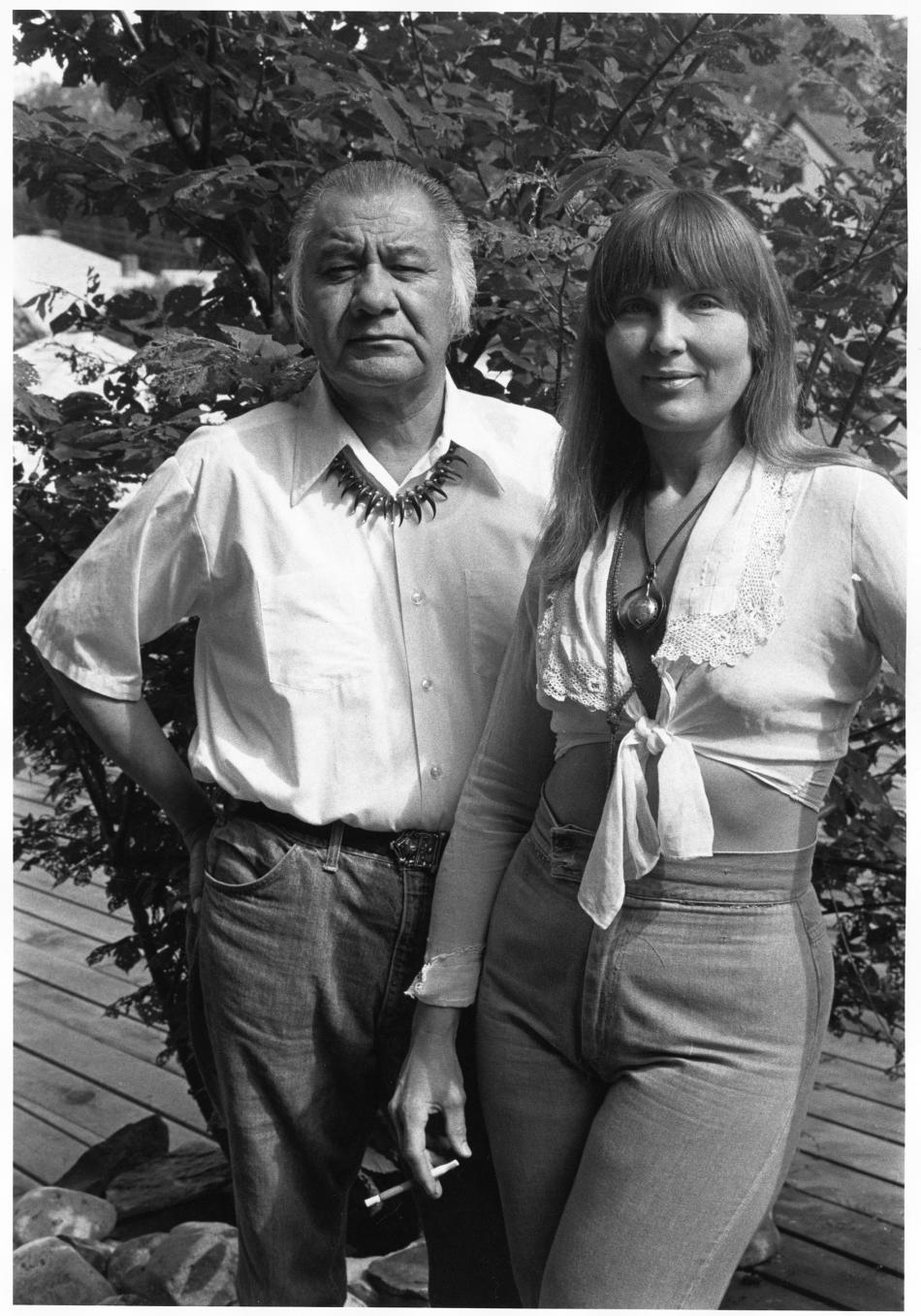A black and white photograph shows a man and woman standing together, from the knees up. The man stands on the left and the woman on the right. The man has long greying hair pulled back and wears a white buttown down shirt tucked into dark pants. The woman has long dark hair with bangs and wears a white shirt tied at the chest and denim pants. The both look at the camera. The man's expression is neutral while the woman has a slight smile. Behind them are the leaves of a tree and a wood deck.