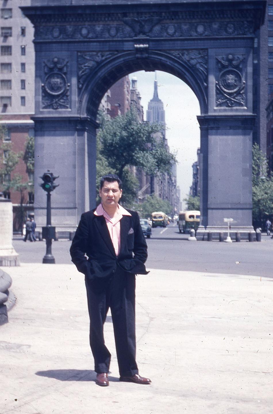 A man with dark brown cropped hair stands with his hands in the pockets of a black suit. He is shown in full-length, standing on pavement with a monumental arch in the distance on a street behind him. There are cars and trucks driving under the arch. He is squinting, looking at the camera. He wears a red and white checkered collared shirt under the suit with a purple pocket square and brown leather shoes.